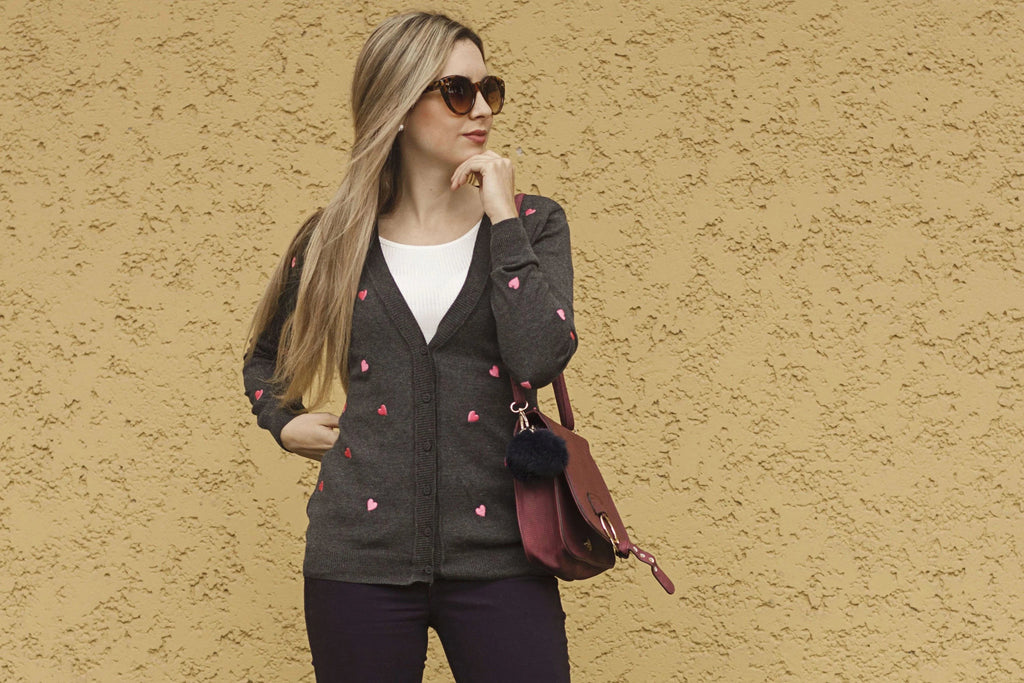 Mujer con suéter gris con corazones rosas, gafas de sol y bolso burdeos frente a pared amarilla