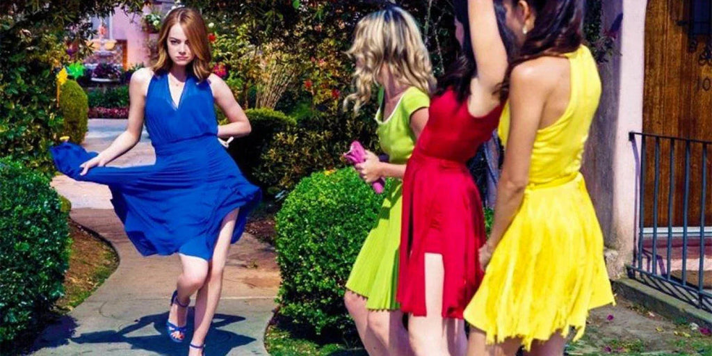 Cuatro mujeres jóvenes en vestido corto de colores azul, verde, rojo y amarillo caminando en un jardín con arbustos y flores