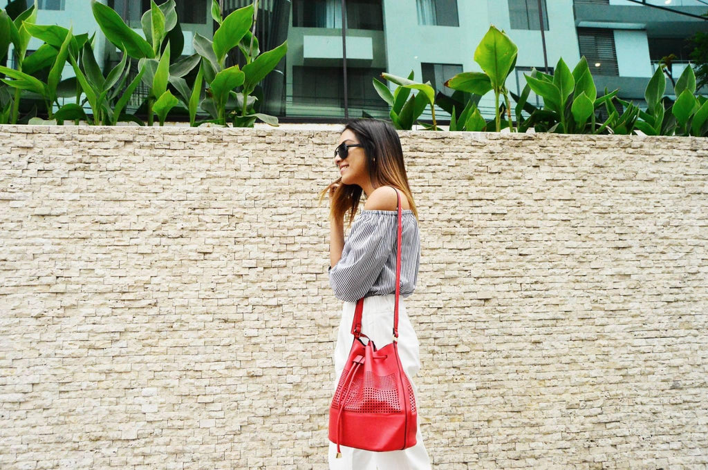 Mujer sonriente con blusa de rayas, pantalón blanco y bolso rojo cruzado frente a muro de piedra y plantas verdes