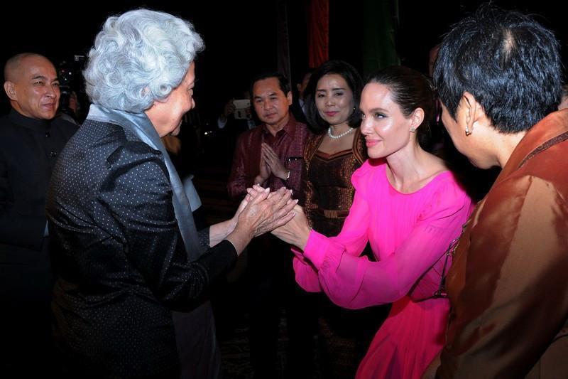 Mujer con vestido rosa brillante saludando a mujer mayor con cabello blanco en evento formal con público observando