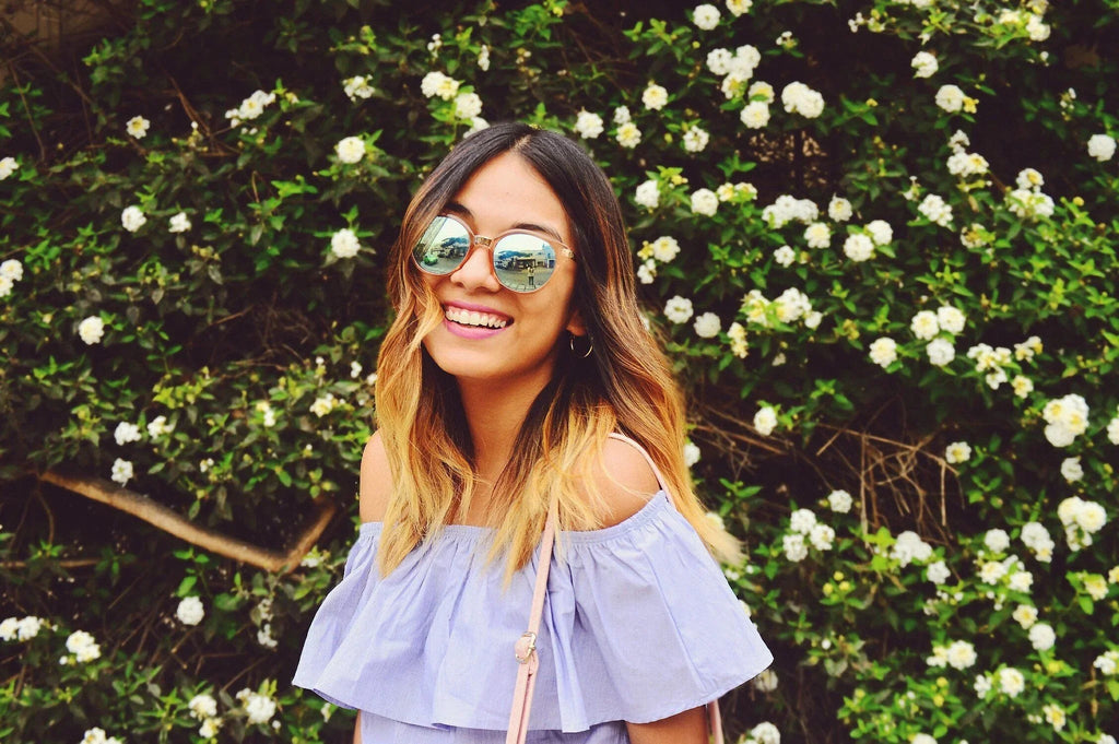 mujer sonriente con gafas de sol redondas, cabello ombré, y blusa azul claro, frente a arbusto con flores blancas