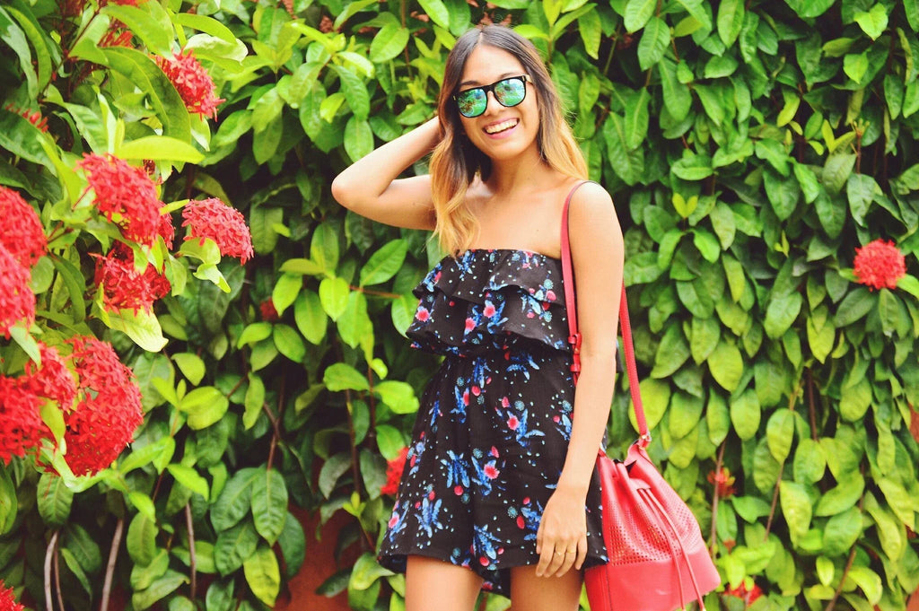 Mujer sonriente con vestido floral negro y gafas de sol frente a arbustos verdes con flores rojas