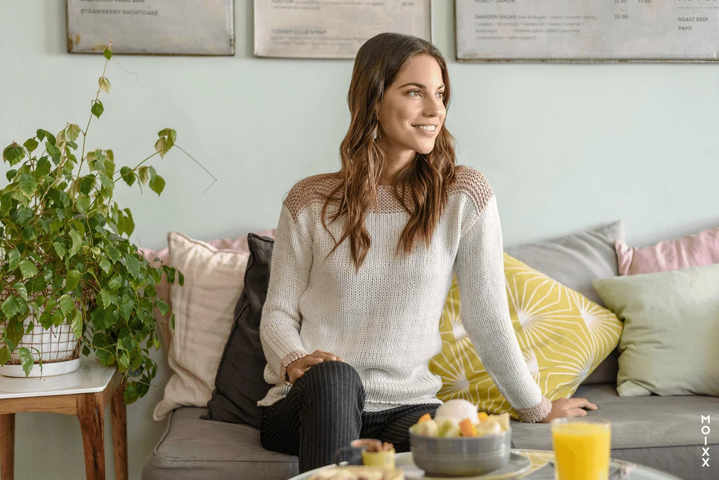 Mujer sonriendo con suéter blanco sentada en sofá con cojines coloridos y mesa con desayuno y planta