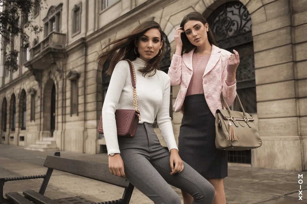 Dos mujeres con estilo urbano posan con ropa elegante y bolsos frente a edificio clásico en la ciudad