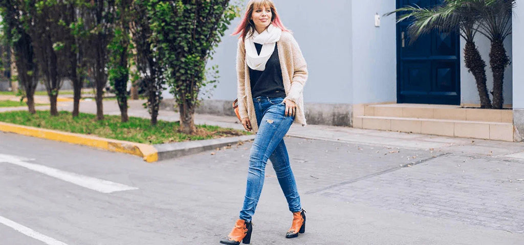 Mujer joven caminando en calle urbana con suéter beige, bufanda blanca y jeans ajustados