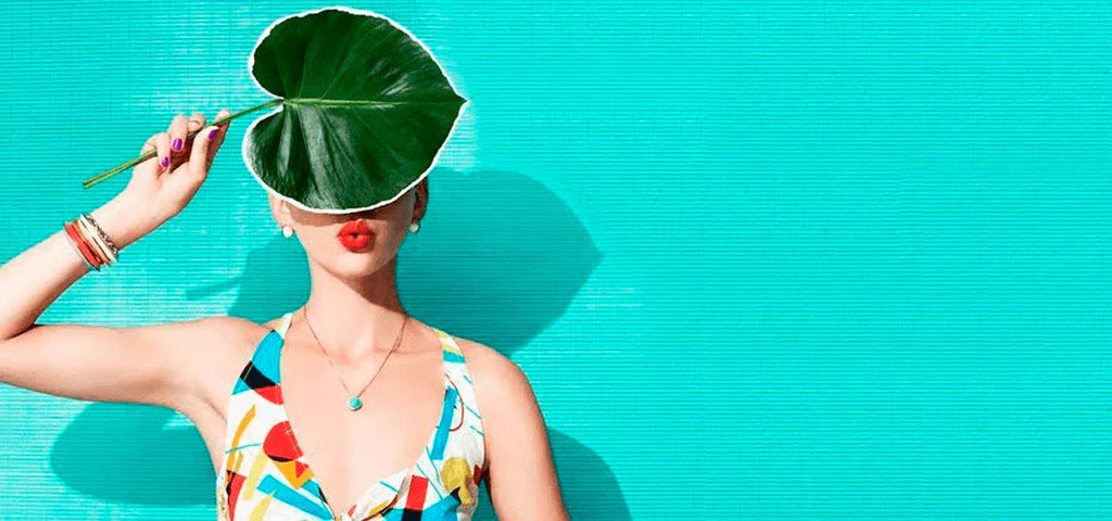 Mujer con vestido colorido y labios rojos cubriendo su rostro con una hoja grande verde frente a pared azul