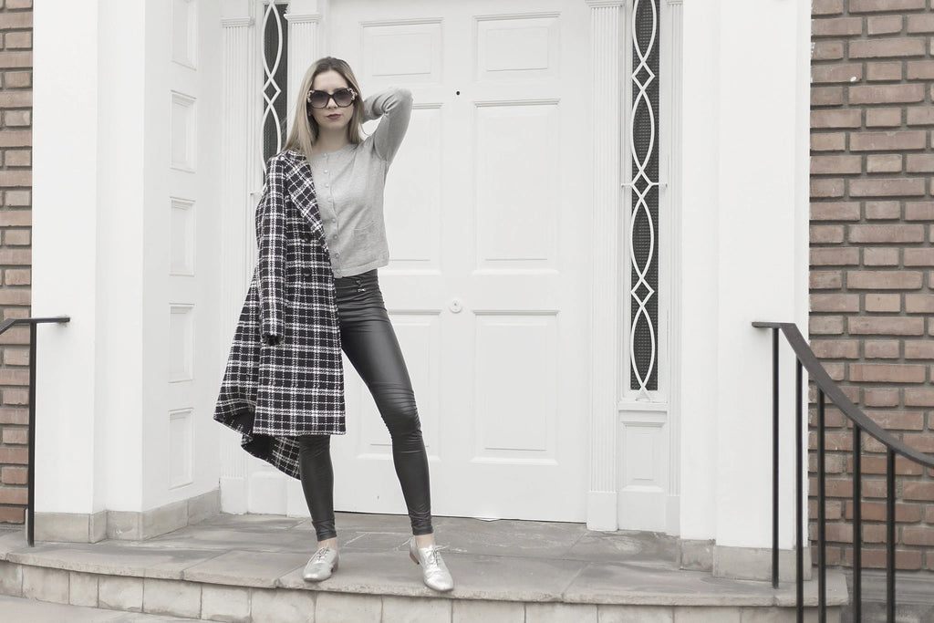 Mujer con gafas de sol y abrigo cuadros negro y blanco sobre el hombro, posando frente a puerta blanca y pared ladrillo