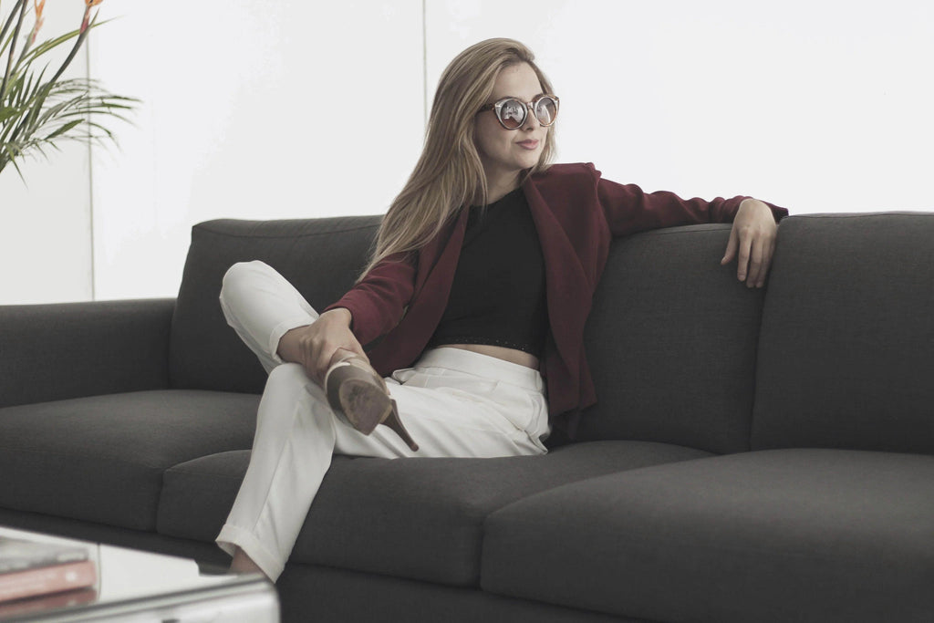 Mujer con chaqueta burdeos, pantalones blancos y gafas de sol sentada en sofá gris moderno
