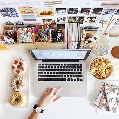 Mesa de trabajo creativa con laptop, marcadores, libros, donuts y palomitas, manos en teclado