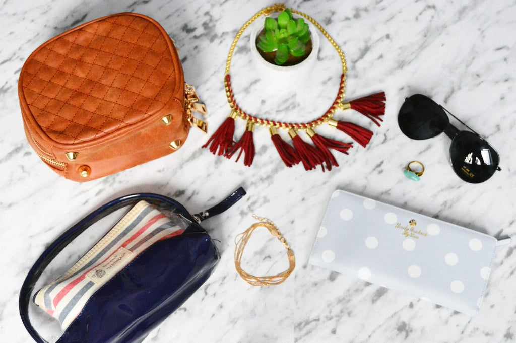 Bolso marrón acolchado, collar con flecos rojos, gafas de sol negras y cartera azul con lunares blancos sobre mármol