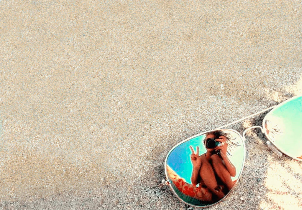 Gafas de sol en la arena con reflejo de una persona haciendo signo de paz y tomando foto en la lente