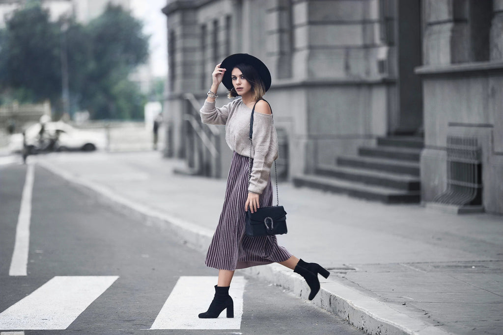 Mujer con sombrero negro, suéter beige y falda plisada caminando sobre paso de cebra urbano con botas negras