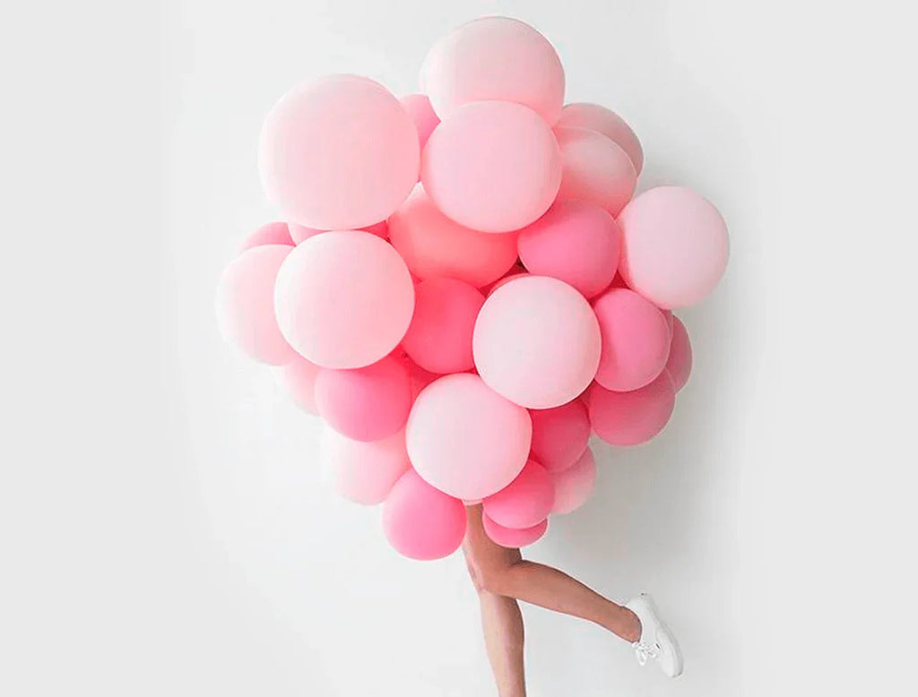Persona cubierta con gran racimo de globos rosados y blancos, fondo blanco minimalista