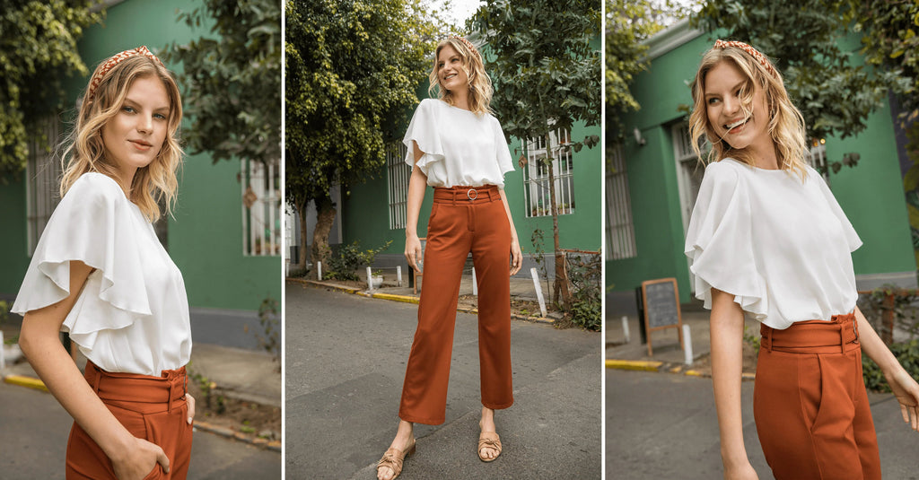 Mujer con blusa blanca de manga capa y pantalones naranjas de tiro alto posando en la calle verde