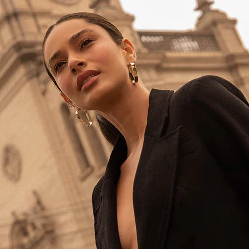 Mujer con chaqueta negra y pendientes dorados en primer plano frente a arquitectura histórica de piedra