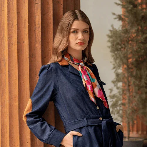 Mujer con vestido azul con parches marrones y pañuelo colorido posando junto a columnas de madera