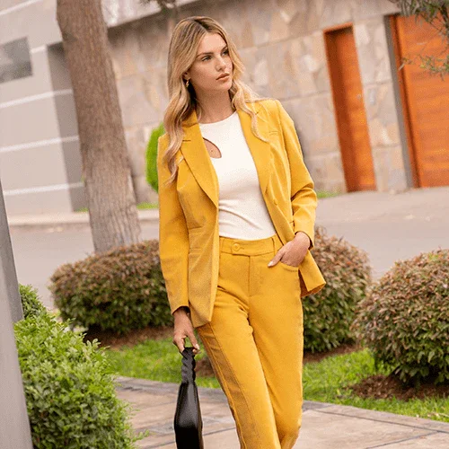 Mujer elegante con traje amarillo, blusa blanca y bolso negro caminando en barrio urbano con arbustos