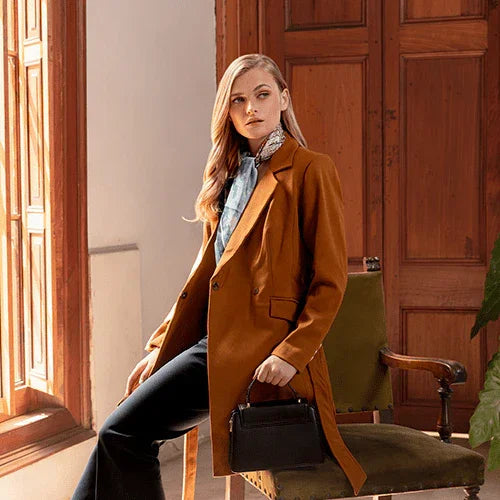 Mujer con abrigo marrón y pantalones negros sentada junto a ventana en sala con muebles de madera