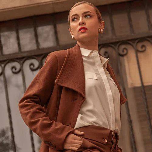 Mujer joven con chaqueta marrón y camisa blanca posando frente a una reja de hierro decorativa