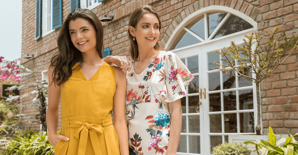 Dos mujeres sonrientes con vestidos de verano, una amarilla y otra blanca con flores, frente a casa de ladrillo