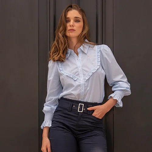 Mujer con blusa celeste de volantes y pantalones negros con cinturón posando frente a pared gris oscuro