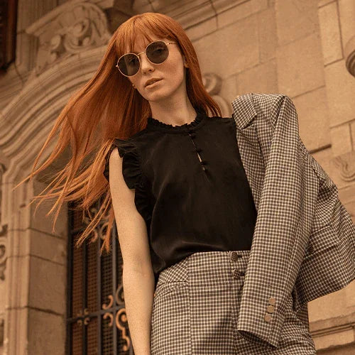 Mujer pelirroja con gafas de sol, blusa negra y traje de cuadros frente a edificio histórico