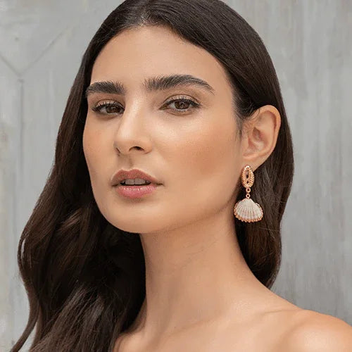 Mujer con piel radiante y cabello oscuro lleva pendientes de concha beige y dorados en fondo neutro