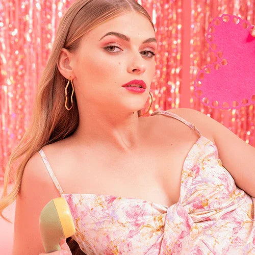 Mujer joven con vestido floral rosa y maquillaje elegante, fondo rosa brillante, sosteniendo teléfono retro