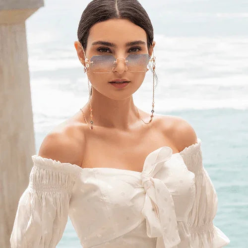 Mujer con gafas de sol degradadas y blusa blanca de hombros descubiertos frente al mar