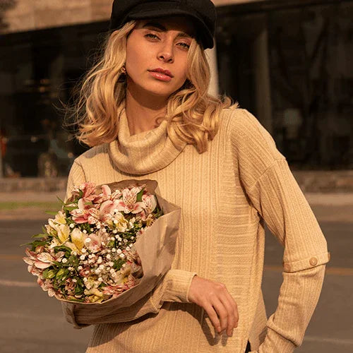 Mujer rubia con suéter beige y boina negra sosteniendo ramo de flores en exterior urbano al atardecer