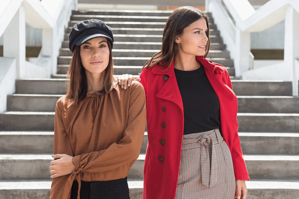 Dos mujeres posando en escaleras urbanas, una con blusa marrón y boina negra, otra con abrigo rojo y falda de cuadros