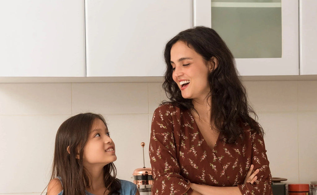 Mujer y niña sonrientes conversando en cocina con cafetera de prensa francesa de cobre