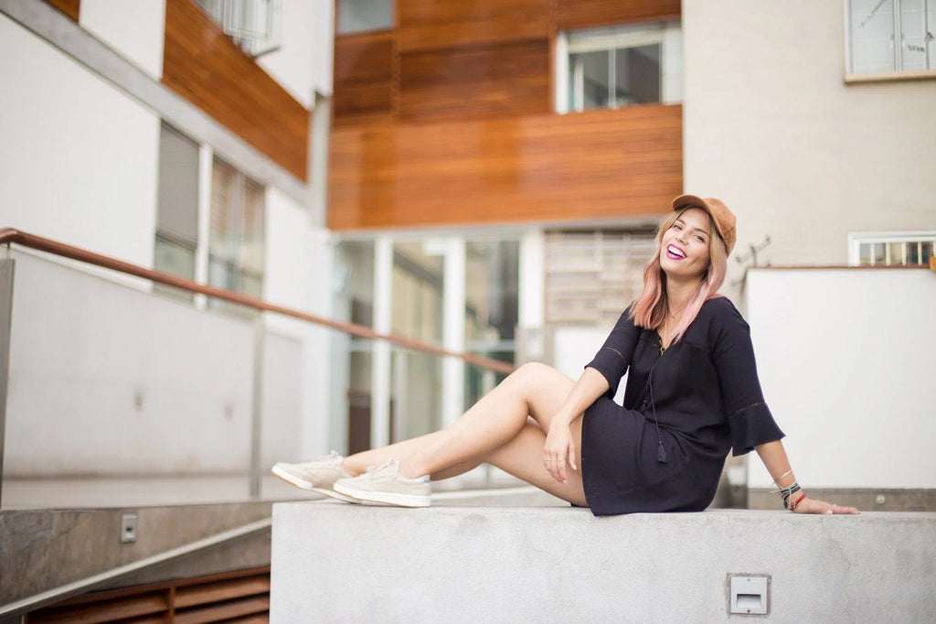 Mujer joven sonriendo con vestido negro y gorra marrón sentada en muro gris en zona urbana moderna