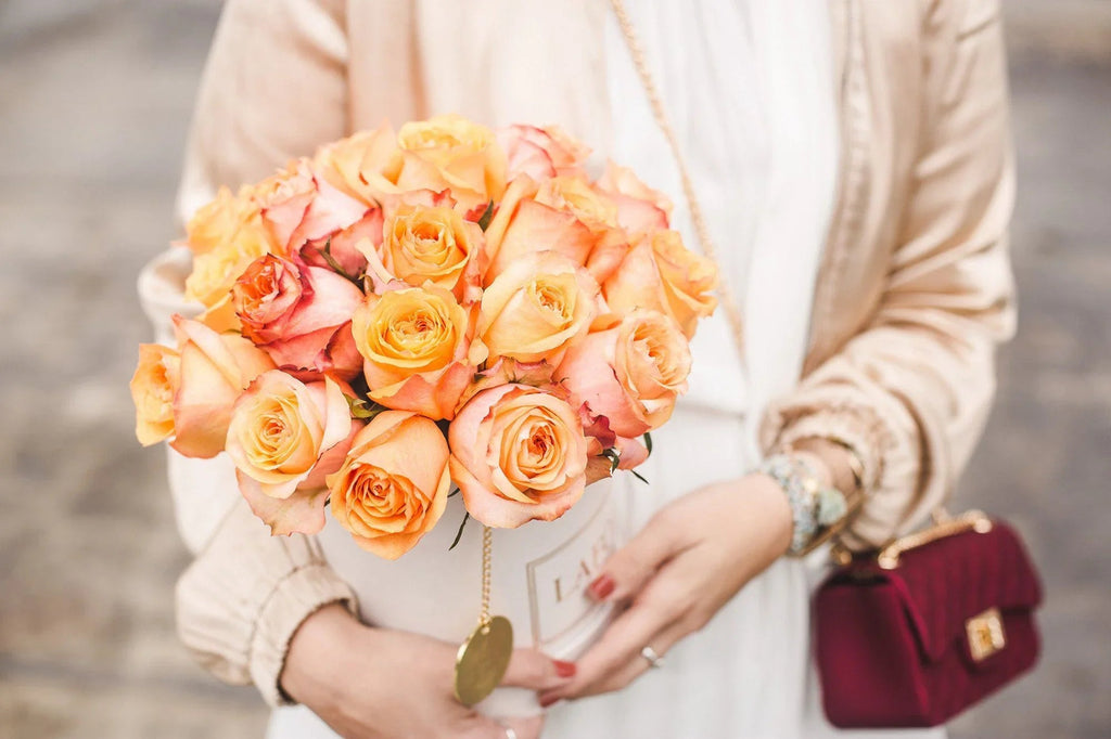 Ramo de rosas naranjas y durazno en caja blanca sostenida por mujer con bolso y ropa beige