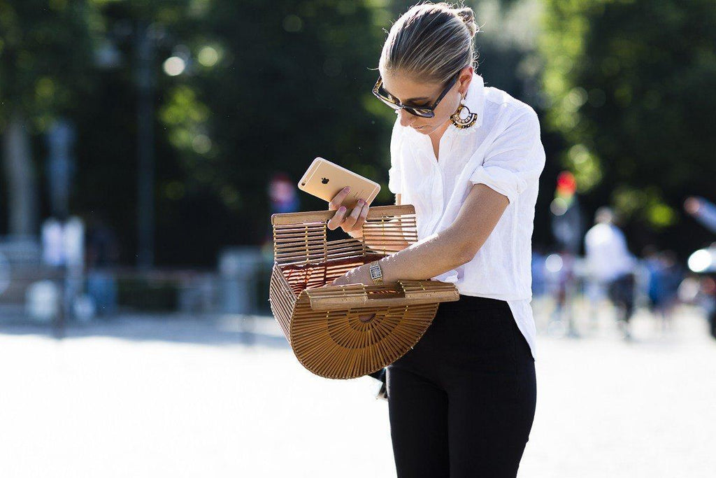 Mujer con camisa blanca y gafas de sol, revisando bolso de madera y sosteniendo smartphone dorado al aire libre