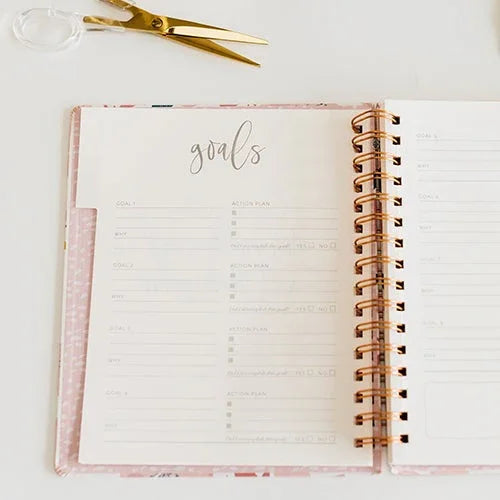 Agenda abierto con planificador de metas y tijeras doradas sobre fondo blanco