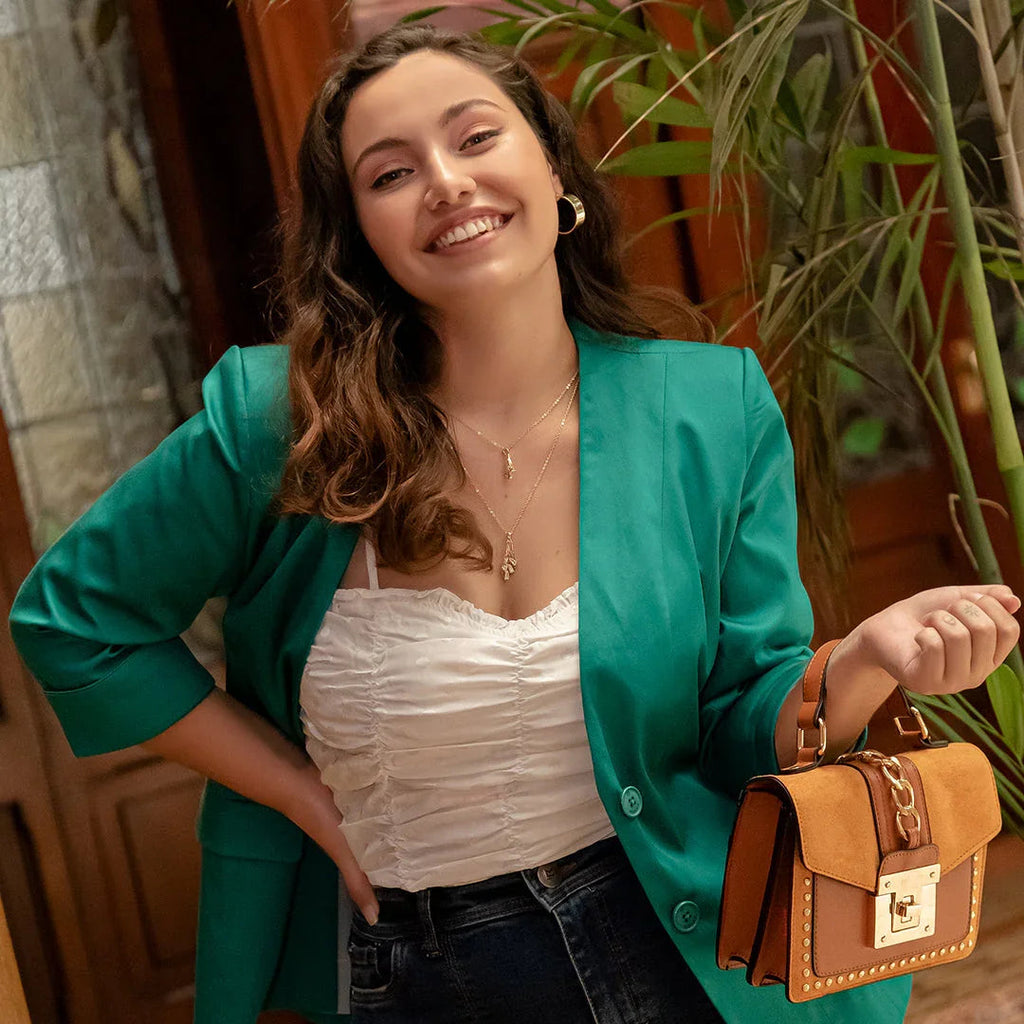 Mujer sonriente con blazer verde y blusa blanca, sosteniendo bolso marrón en interior con plantas