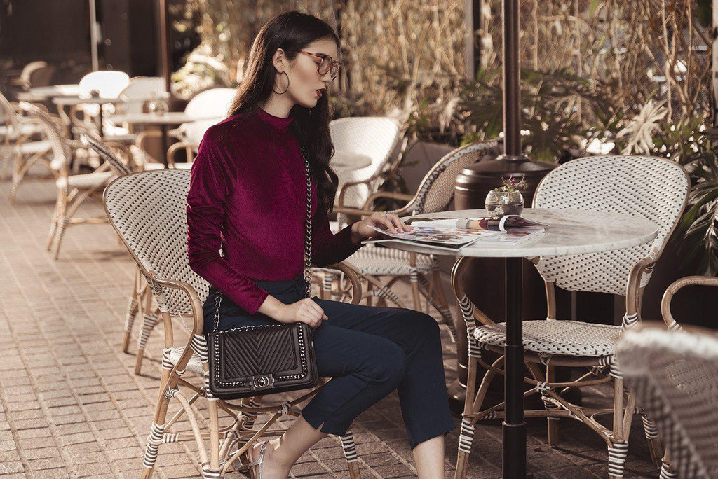 Mujer con blusa vino, pantalones oscuros y gafas leyendo revista en terraza con mesa y sillas de mimbre
