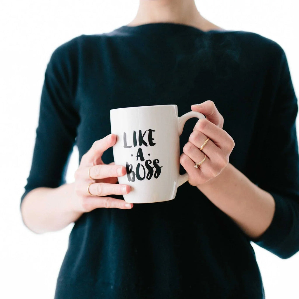 Persona con suéter negro sostiene taza blanca con texto Like a Boss y vapor de bebida