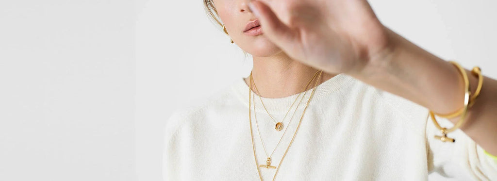 Mujer con collar dorado en capas y suéter blanco levantando la mano frente a fondo blanco