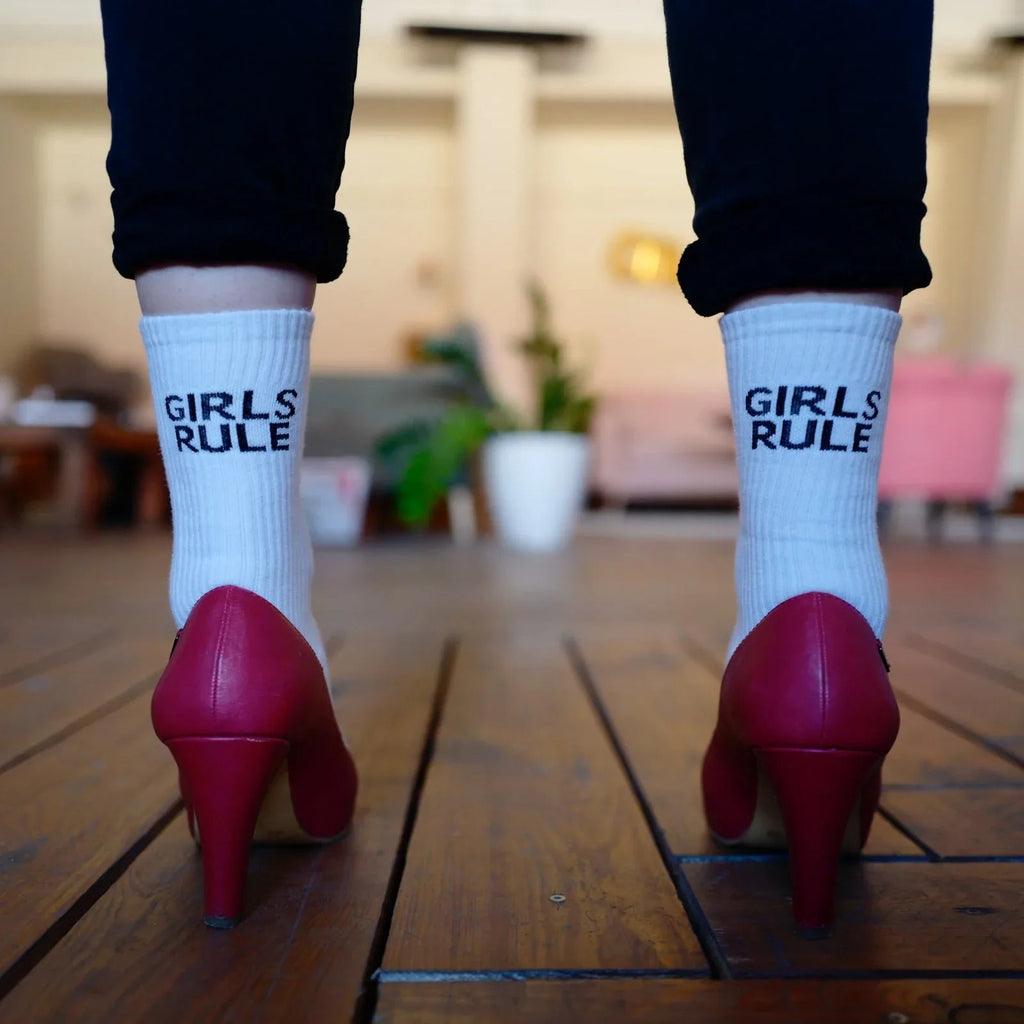Pies con calcetas blancas con texto Girls Rule y zapatos de tacón rojo sobre piso de madera