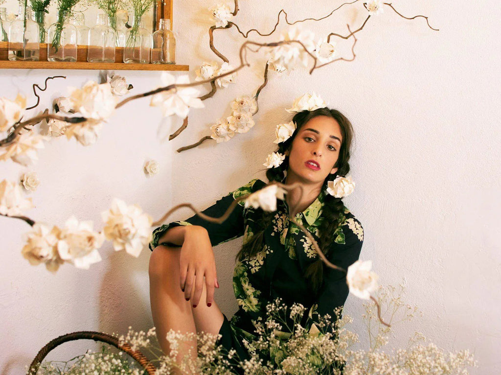 Mujer con trenzas y flores blancas en el cabello, en ambiente floral y vestido con estampado verde y negro