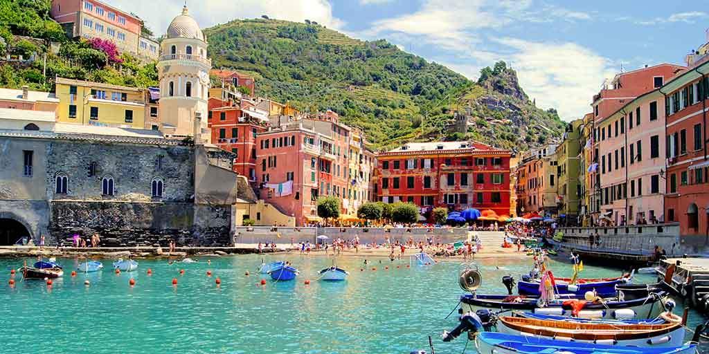 Puerto con barcas y edificios coloridos frente a una playa con bañistas bajo cerros verdes en Italia