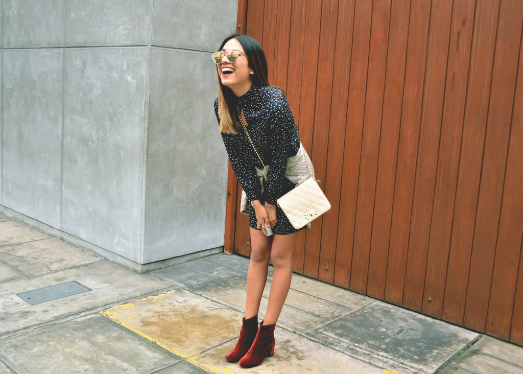 Mujer con vestido negro de lunares, botas rojas y bolso beige sonriendo en calle con pared de madera y cemento