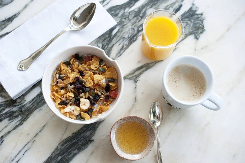 Desayuno saludable con cereal mixto en bol, zumo de naranja, café con leche y miel sobre mesa de mármol