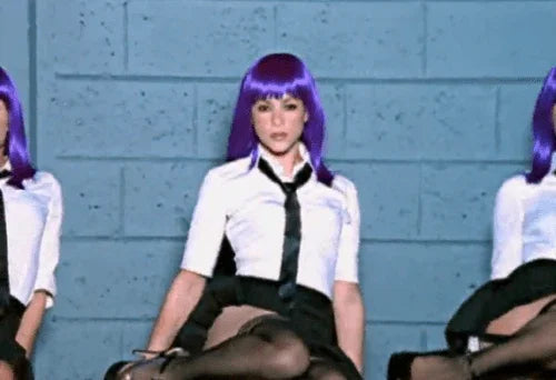 Mujer con peluca morada y uniforme escolar blanco y negro sentada frente a fondo azul