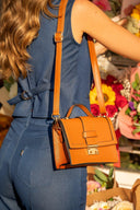 Bolso de cuero color caramelo con hebilla dorada, mujer con outfit azul y flores al fondo