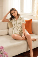 Mujer sentada en sofá beige con pijama corto blanco con estampado verde y cojín naranja en sala luminosa