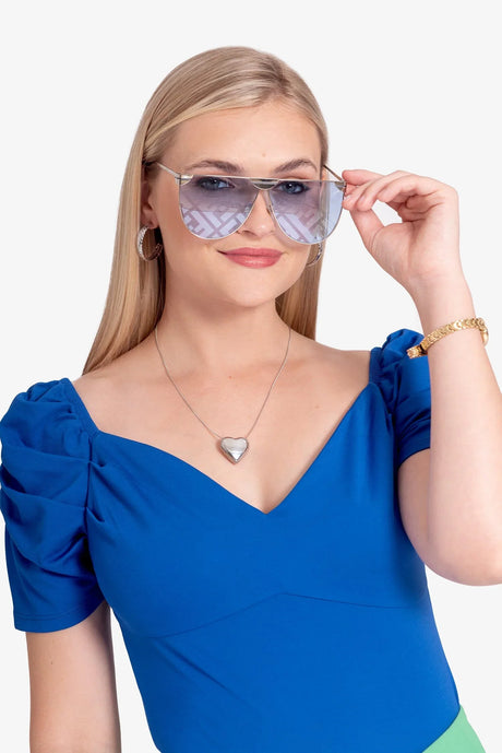 Mujer rubia con blusa azul y gafas de sol reflectantes, joyería de plata y pulsera dorada